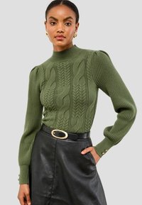 Grüner Strickpullover mit Kabelmustern, gerippten Bündchen und hohem Kragen. Kombiniert mit einem schwarzen Lederrock und einem Gürtel mit goldener Schnalle.