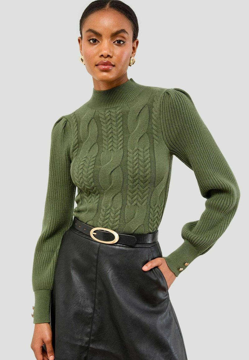 Grüner Strickpullover mit Kabelmustern, gerippten Bündchen und hohem Kragen. Kombiniert mit einem schwarzen Lederrock und einem Gürtel mit goldener Schnalle.