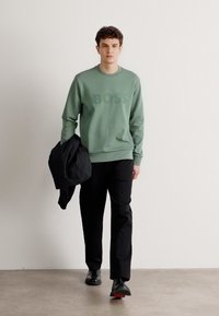Felpa verde con logo "BOSS", pantaloni neri e giacca nera tenuta in mano. Tessuto morbido, vestibilità casual, modello che cammina su uno sfondo semplice.