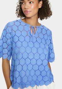 Top in pizzo blu chiaro con motivo floreale, maniche corte e dettaglio a fiocco al collo. Orlo scallop e texture intricata in tutto il capo.