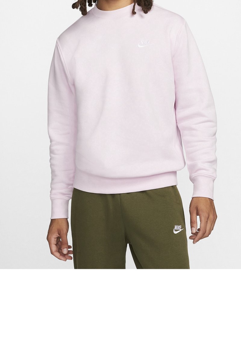 Homme portant un sweat-shirt Nike rose clair et un pantalon de survêtement Nike vert olive, debout devant un fond blanc uni.