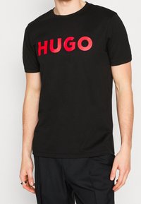 Musta puuvillapaita, jossa on rohkea punainen "HUGO"-logo rintamuksessa, lyhyet hihat ja tavallinen pyöreä kaula-aukko.