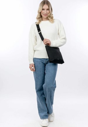 Svart axelväska med justerbar rem, rektangulär form, slät textur och silverdetaljer. Modell bär en vit tröja och blå jeans.