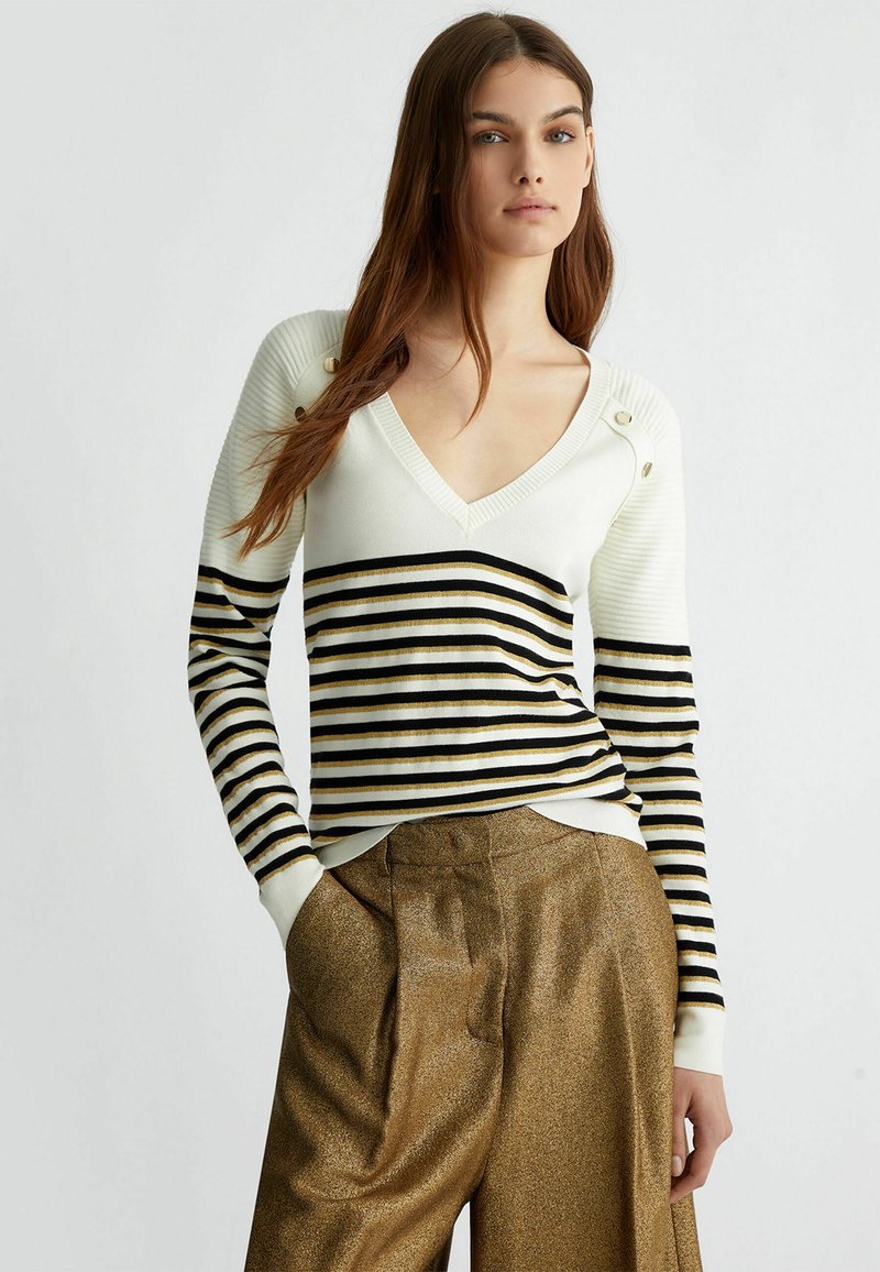 Maglione lavorato a maglia di colore crema con strisce nere e dorate, dotato di scollatura a V e dettagli con bottoni sulle spalle. Texture morbida e design aderente.