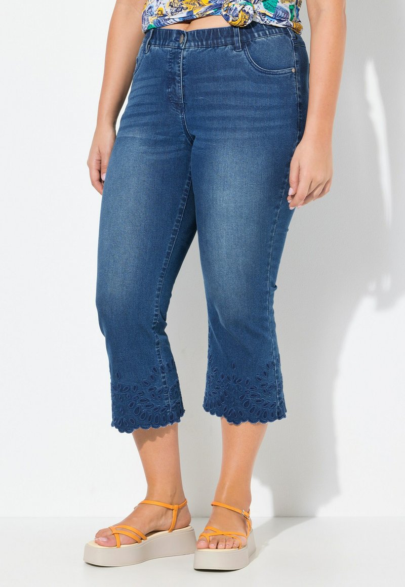 Pantalones capri de mezclilla en azul oscuro con un dobladillo bordado intrincado, que presentan un ajuste ceñido y una cintura de altura media, combinados con sandalias de plataforma color marrón.