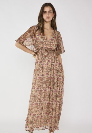 Giovane donna che indossa un lungo abito floreale con maniche ampie, in piedi con la mano sul fianco davanti a uno sfondo chiaro uniforme.