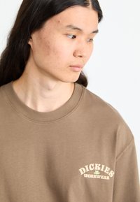 Brun kortermet sweatshirt med rund hals. Har en kremfarget logo med teksten "DICKIES WORKWEAR" på venstre brystområde.