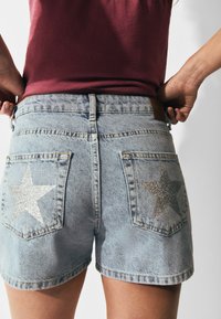Shorts de denim en azul claro con dos bolsillos traseros que presentan patrones de estrellas de brillo plateado. Corte clásico con un dobladillo deshilachado y parche de cuero.