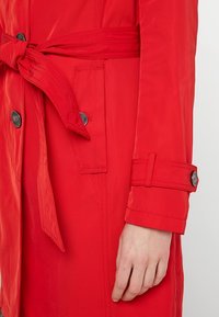 Gabardina roja con cinturón en la cintura, puños abotonados, dos bolsillos frontales y una textura suave. La tela tiene un ligero brillo.