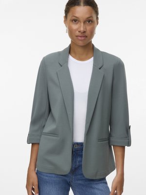 Blazer gris clair avec un design ajusté, des revers à pointes et des manches retroussées. Porté sur une chemise blanche et un jean bleu, avec des poches latérales.