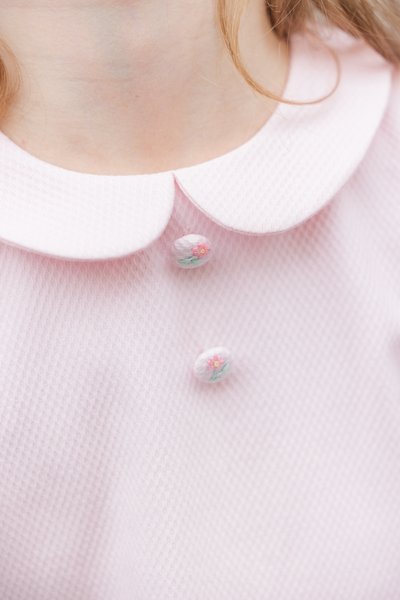 Ροζ ανοιχτόχρωμο ύφασμα με υφή, με στρογγυλεμένο γιακά, που έχει δύο διακοσμητικά κουμπιά με κεντήματα λουλουδιών.