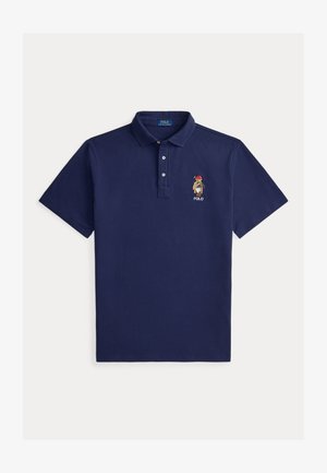 Temno modra polo majica s kratkimi rokavi, ovratnikom, tremi gumbi in vezenim medvedom, ki nosi rdečo kapo in drži golf palico nad napisom "POLO".