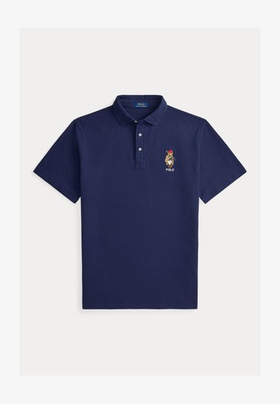 Μπλε σκούρο κοντομάνικο πόλο μπλουζάκι με γιακά, τρία κουμπιά και κεντημένη αρκούδα που φοράει κόκκινο καπέλο και κρατάει ένα γκολφ κλαμπ πάνω από το κείμενο "POLO".