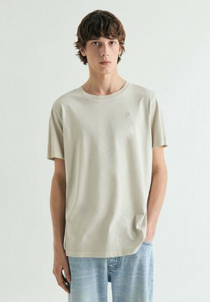 Camiseta de algodón color beige claro con cuello redondo, mangas cortas y un logo sutil en el pecho, combinada con jeans de mezclilla azul claro.