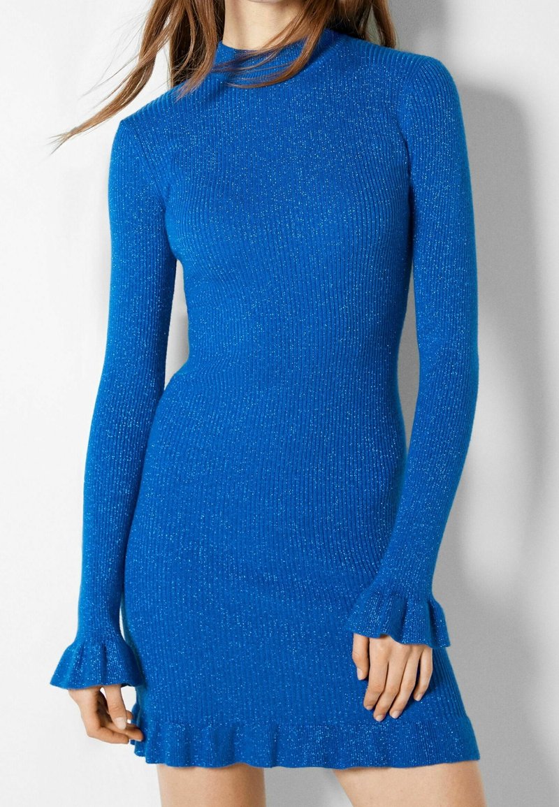 Robe tricotée côtelée bleue avec manches longues, manches évasées et ourlet volanté. Le matériau a une texture scintillante. Design à col haut.