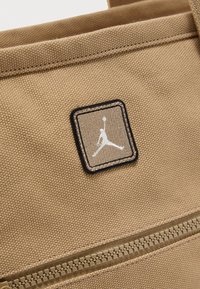 Gros plan d'un sac en tissu beige avec un patch rectangulaire cousu présentant la silhouette blanche d'un joueur de basketball et une fermeture éclair beige en dessous.