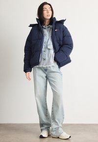 Ναυτικό μπούφασο μπουφάν με ψηλό γιακά, συνδυασμένο με ανοιχτό τζιν πουκάμισο και φαρδιά ανοιχτόχρωμα τζιν παντελόνια, φορεμένα με λευκά αθλητικά παπούτσια.