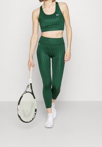 Zielony sportowy biustonosz i dopasowane legginsy z fakturowanym wzorem. Modelka trzyma rakietę tenisową; na nogach białe buty sportowe.