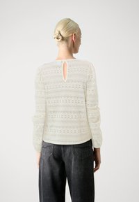 Blouse en dentelle blanche à manches longues, avec un détail de dos en trou de serrure et des motifs délicats. Associée à un jean noir fuselé.