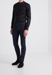 Maglia nera a maniche lunghe con collo alto e bottoni sul davanti, abbinata a jeans slim fit blu scuro e stivaletti in suede neri.