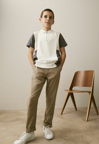 Polo à manches courtes avec une fermeture éclair en crème et gris, associé à un pantalon beige et des baskets blanches. Chaise en bois en arrière-plan.