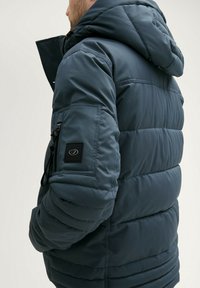 Mornarska podložena jakna s kapuco, ki ima žep z zadrgo na rokavu in teksturirano površino. S prilagodljivimi vrvico.