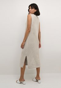 Ärmelloses Strickkleid in hellbeige mit offenem Muster, seitlichem Schlitz und geripptem Saum. Kombiniert mit weißen Blockabsatzsandalen.
