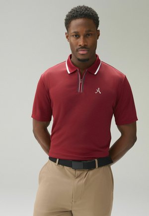 Mann mit kurzen lockigen Haaren, der ein kastanienbraunes Poloshirt und beige Hosen mit schwarzem Gürtel trägt, steht mit Händen hinter dem Rücken.