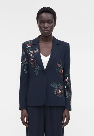 Γυναίκα που φοράει ένα σκούρο μπλε σακάκι με πολύχρωμη κεντητή ανθική διακόσμηση στον έναν ώμο, στο μανίκι και μπροστά, συνδυασμένο με παντελόνια σε ίδιο σκούρο μπλε χρώμα.