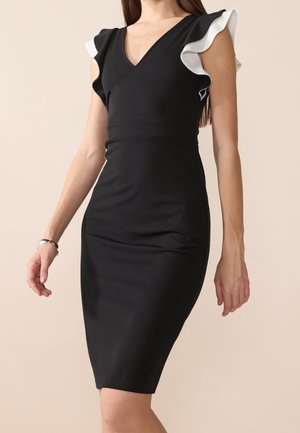 Dopasowana czarna sukienka do kolan z białymi falbanianymi krótkimi rękawami i głębokim dekoltem w serek, noszona przez osobę na jednolitym beżowym tle.