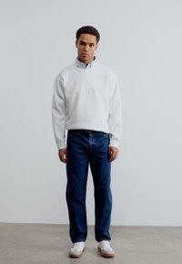 Λευκό φούτερ με υψηλό γιακά, φορεμένο πάνω από ένα γαλάζιο πουκάμισο, συνδυασμένο με μπλε τζιν και λευκά αθλητικά παπούτσια. Μινιμαλιστικός σχεδιασμός.