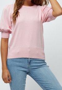 Helles rosa Strickpullover mit rundem Halsausschnitt und puffierten kurzen Ärmeln. Gerippter Saum und Bündchen. Kombiniert mit blauen Jeans.