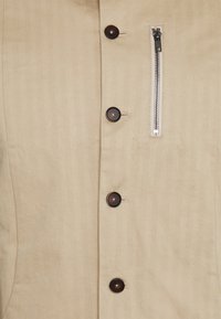 Tan jacket featuring a button-front with dark brown buttons and a zippered pocket on the left side, crafted from smooth fabric.