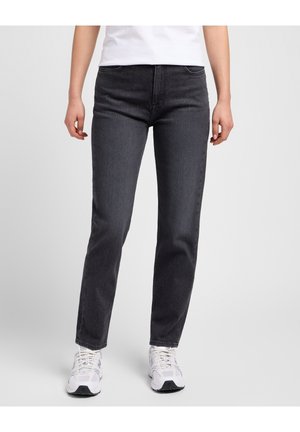 Personne portant un jean noir à jambes droites, des baskets blanches et un haut blanc, se tenant devant un fond uni gris clair.