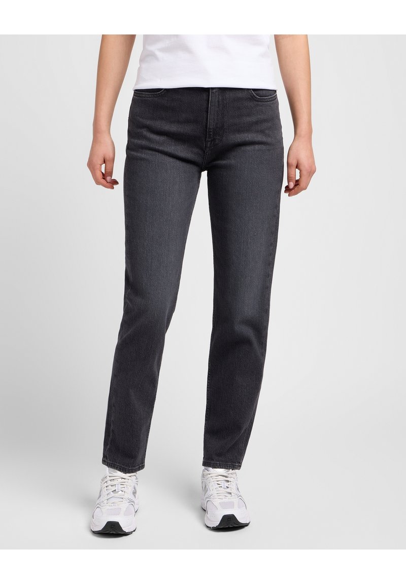 Personne portant un jean noir à jambes droites, des baskets blanches et un haut blanc, se tenant devant un fond uni gris clair.