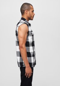 Fekete-fehér kockás, ujjatlan ing, gombos elejével, kockás anyagból készült, fekete nadrággal párosítva, oldalról látható.