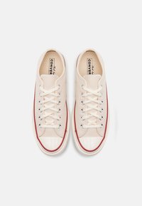 Beige lærersneakers med hvite gummitupper, røde side-striper og runde metalløyehull. Har flate, kremfargede snorer og en teksturert såle.