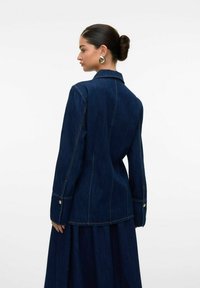 Veste en denim bleu foncé avec une coupe ajustée, dotée de manches longues, d'un col cranté et de coutures visibles. Accents de boutons dorés sur les poignets.