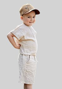 Hellbeigees Leinenoutfit mit einem kurzärmligen Oberteil und knielangen Shorts, mit einem Kordelzugbund und seitlichen Taschen. Braune Baseballmütze mit Design.