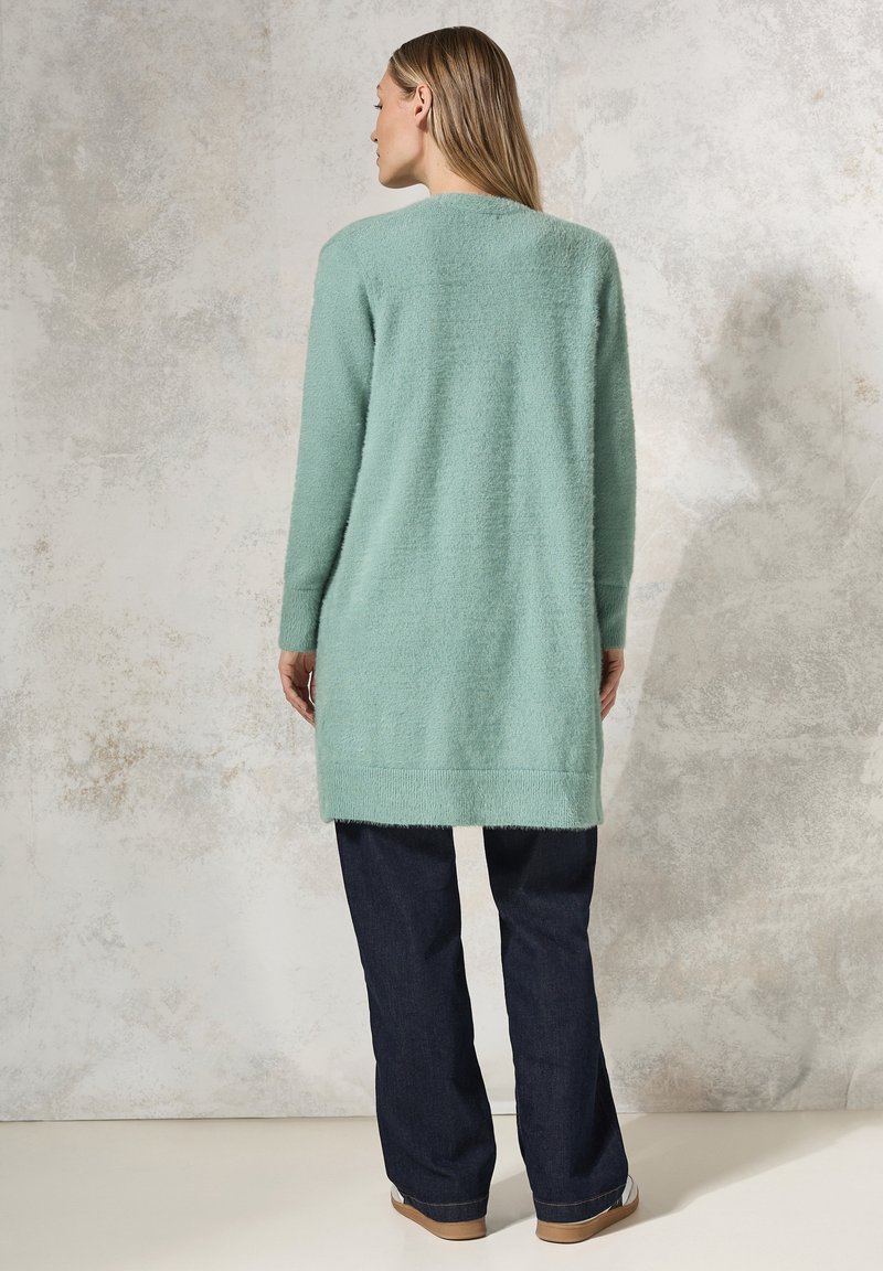 Mintgrüner, langer Cardigan aus weichem, strukturiertem Stoff; kombiniert mit dunkelblauen Hosen; hat einen geraden Schnitt und lange Ärmel.