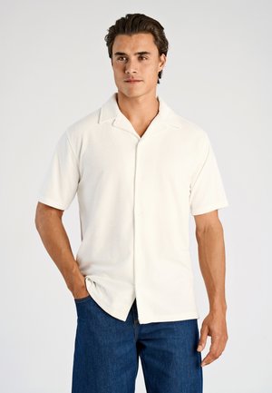 Man met een wit, kort mouwig overhemd met knopen en een kraag, en een blauwe spijkerbroek, staand met één hand in de zak tegen een witte achtergrond.