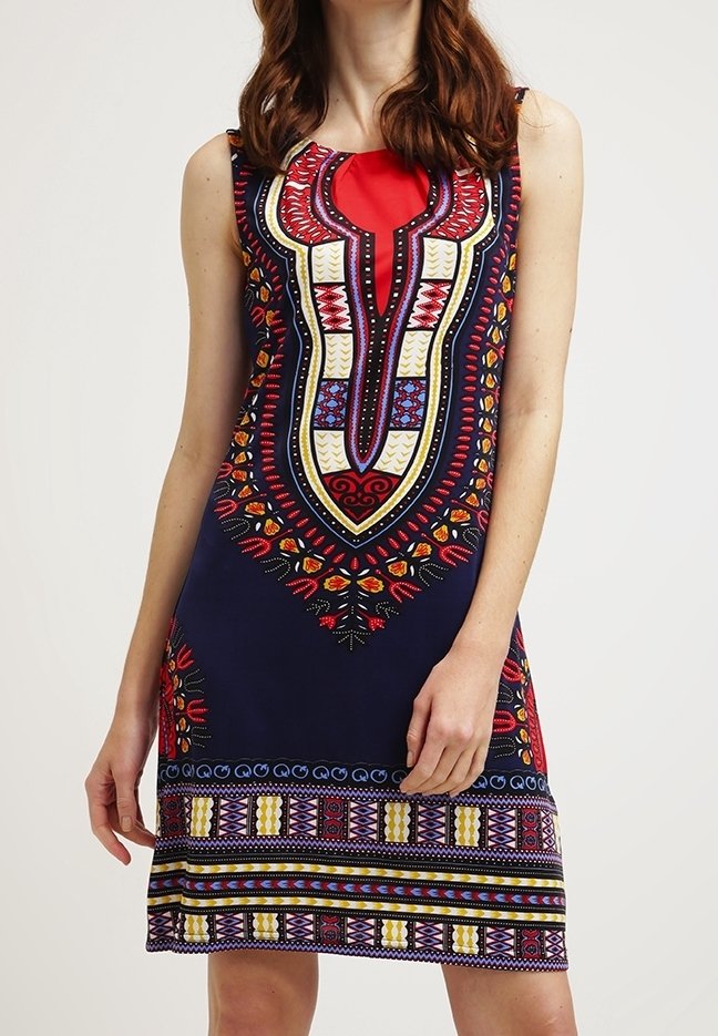 Robe bleu marine sans manches avec des motifs géométriques et floraux africains vibrants en rouge, jaune et bleu, portée par une personne debout aux cheveux ondulés.