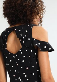 Robe noire avec des pois blancs et roses, épaules dénudées avec des volants, et un détail en trou de serrure au dos. Texture de tissu lisse.