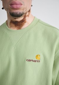 Lys grøn sweatshirt med ribbet halsudskæring. Har et broderet logo i gul og bordeaux på venstre bryst. Blødt, struktureret stof.