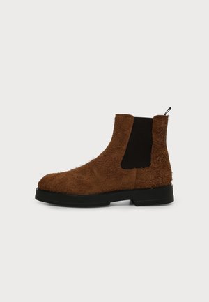 Bottine Chelsea en daim marron avec panneau latéral élastique noir, semelle noire et languette à l'arrière sur fond gris clair uni.