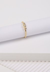 Goldring mit drei klaren Edelsteinen, die in einem eleganten Band gefasst sind. Glatte Textur und glänzende Oberfläche vor einem weißen Hintergrund.