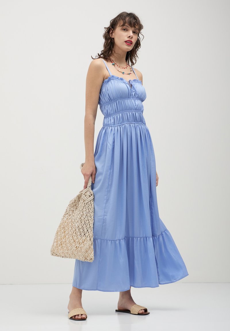 Frau in einem hellblauen ärmellosen Maxikleid, die eine beige gewebte Handtasche hält, Riemchensandalen trägt und eine bunte Perlenkette um den Hals hat.