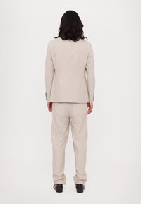 Costume en lin beige comprenant un blazer ajusté et un pantalon ample. Texture lisse avec des détails de couture minimaux et des chaussures noires.