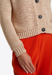 Pletený cardigan v světlé béžové barvě s texturovaným vzorem, zapínání na knoflíky a žebrovanými manžetami, v kombinaci s jasně oranžovými kalhotami.