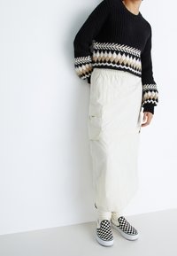 Czarny sweter dziany z teksturowanymi rękawami i geometrycznym wzorem; kremowa spódnica z kieszeniami typu cargo; czarno-białe wsuwane buty w kratę.